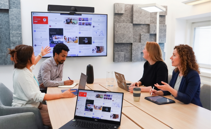 our colleagues in a modern meeting room collaborating and gesturing toward a large interactive wall display, demonstrating human-centric technology in the workplace.